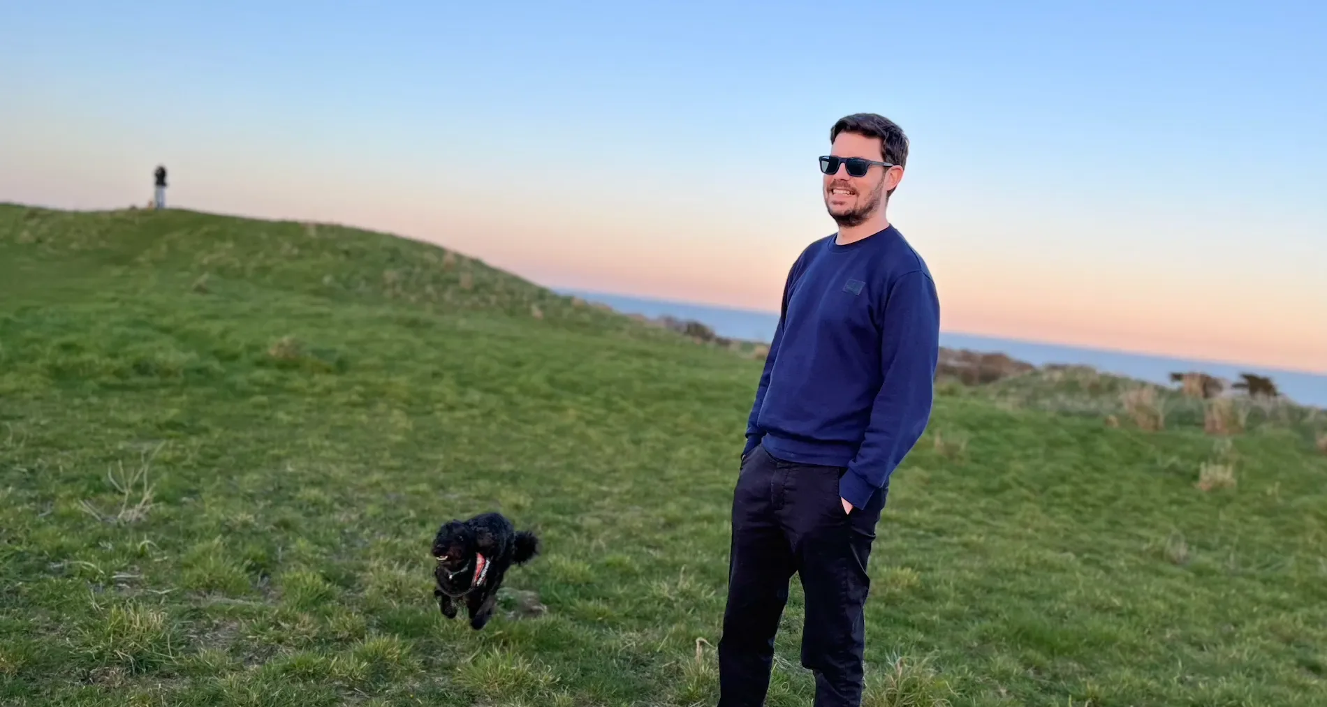 Ian Cumbers standing in a field by the coast, wearing sunglasses and a blue jumper